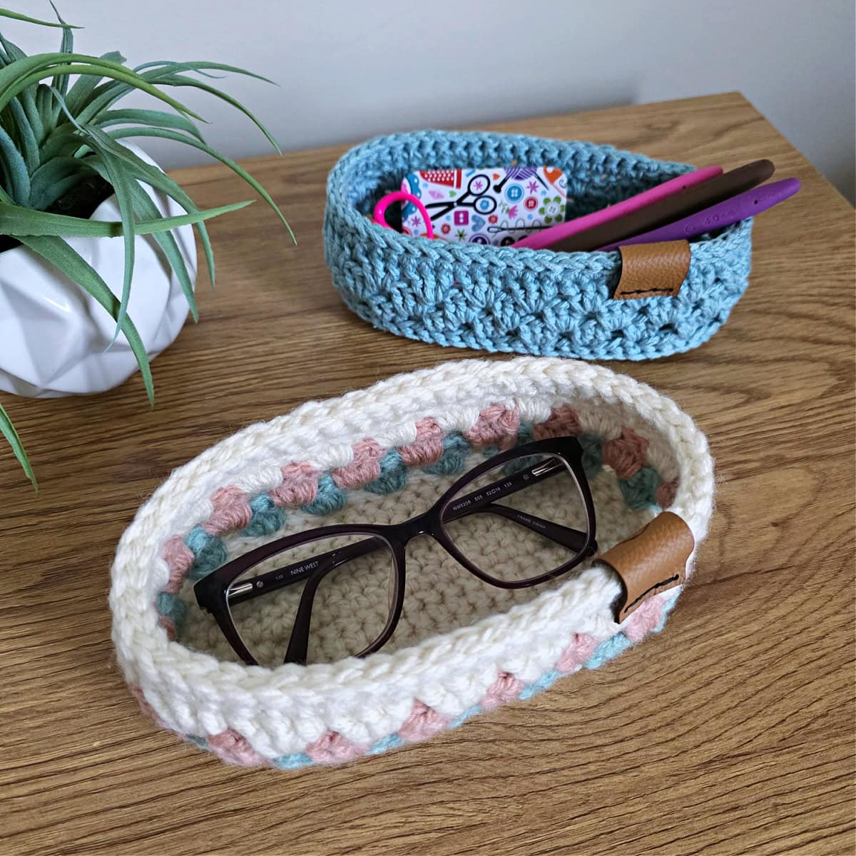 Cream, pink, and aqua crochet glasses holder with eyeglasses in them on a wood desk.