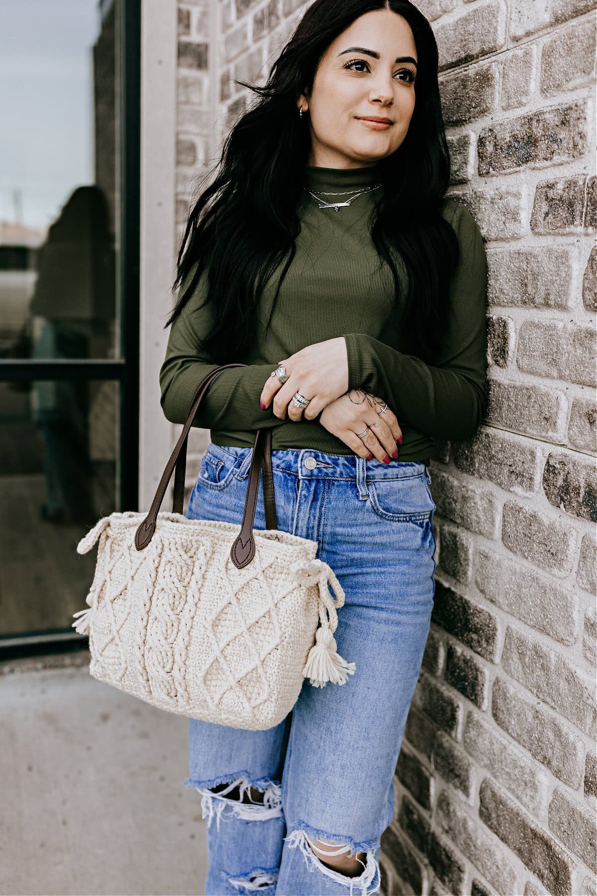 A young woman leaning on a brick wall holding a crochet cabled bag with leather straps.