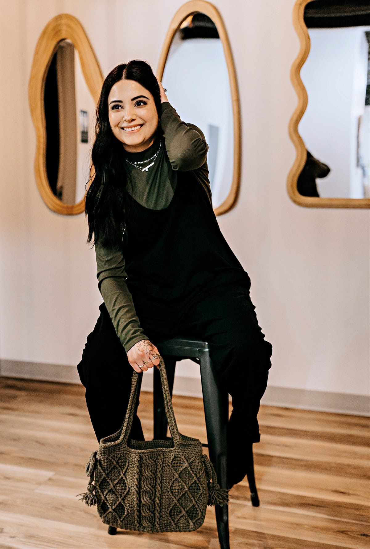 A young girl sitting on a black stool holding a green crochet purse with cables and pleats.