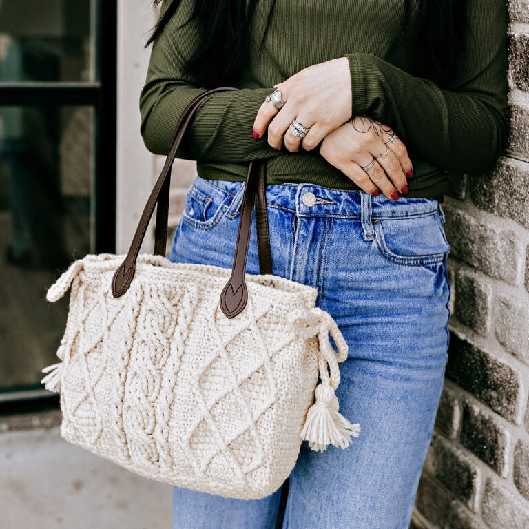 Close-up of a cream color cabled crochet purse with brown leather straps being held by a women in a green top and jeans.