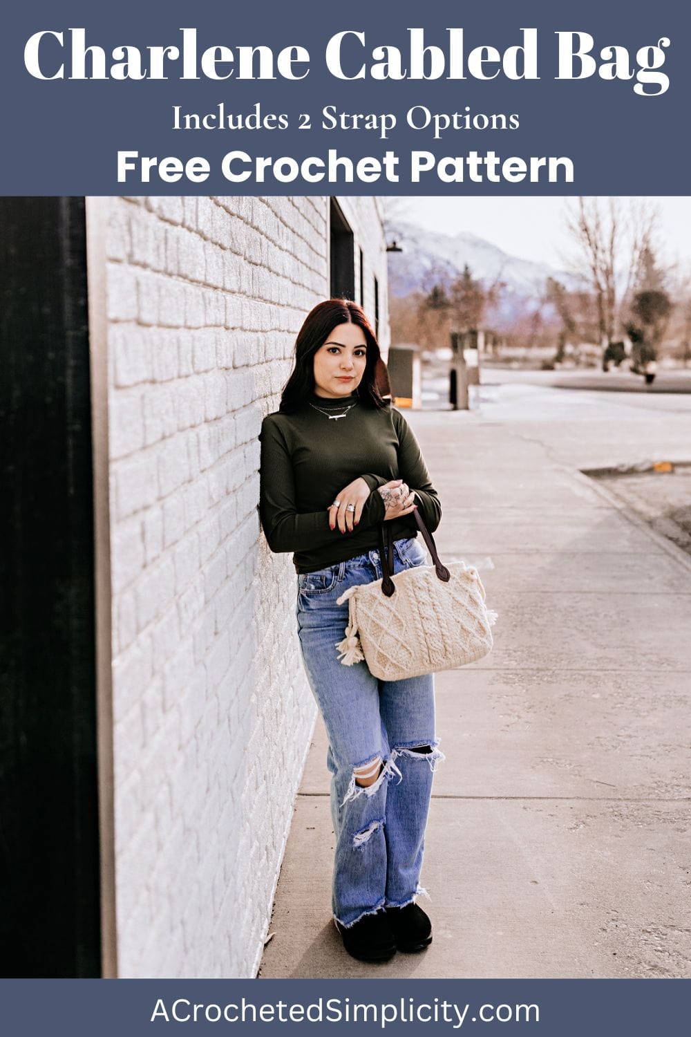 A crochet purse with brown leather straps being held by a women in a green top and jeans who is leaning against a brick building.