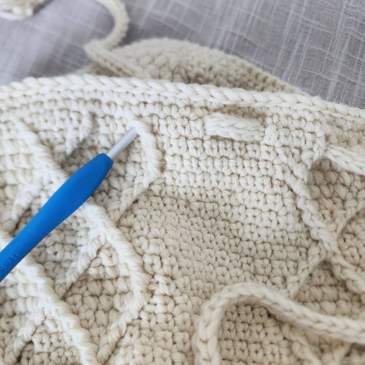 A blue crochet hook inserted into a stitch on the opposite side to pull the icord through another hole.